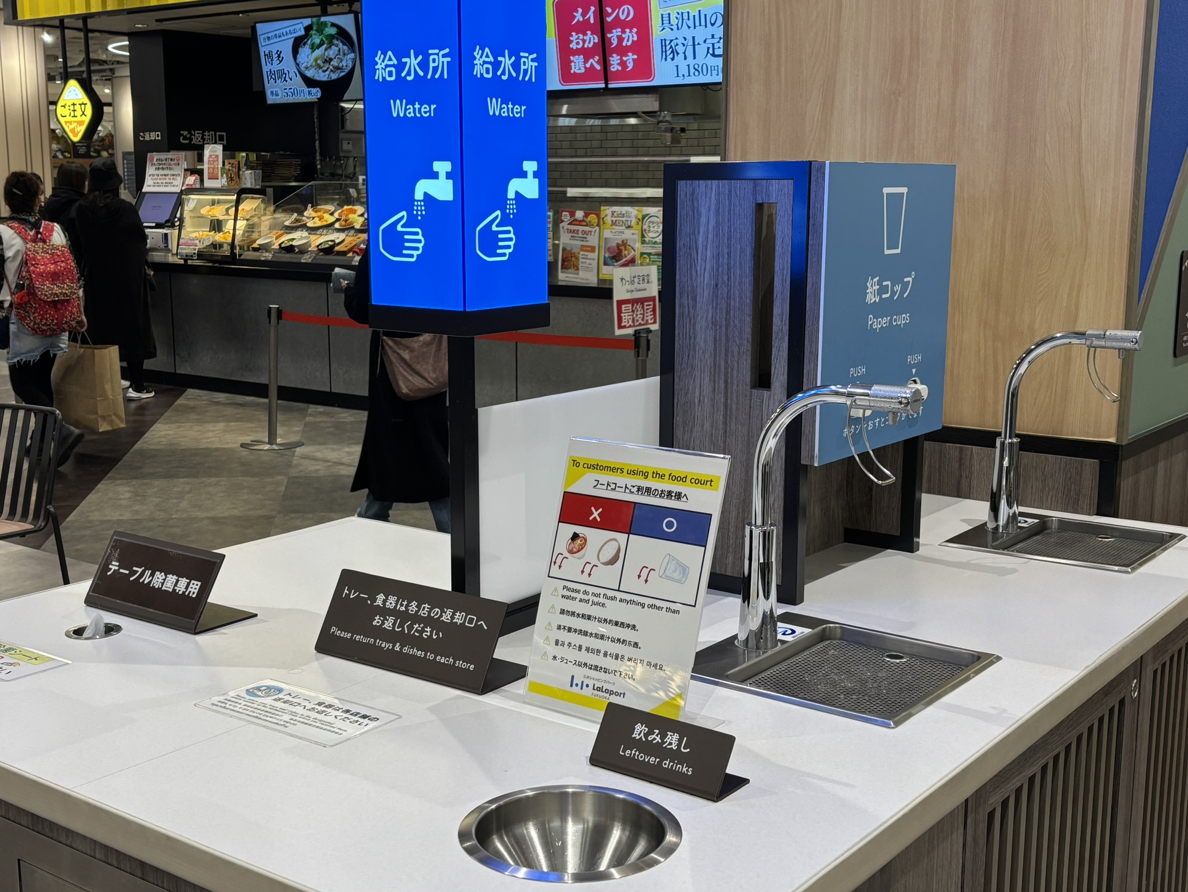 美食廣場的整合式回收與飲水區。設置給水（Water）、紙杯（Paper cups）、以及專門的飲料殘渣傾倒孔（Leftover drinks），將複雜的分類流程化繁為簡，鼓勵使用者自主維持整潔。